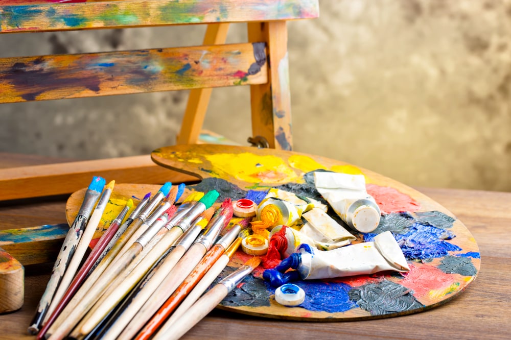 Paleta de pintura de madeira apoiada sobre uma mesa, coberta com tintas coloridas espalhadas em tons de azul, vermelho, amarelo e cinza. Sobre a paleta há tubos de tinta parcialmente amassados e abertos, além de pincéis de diferentes tamanhos com cerdas sujas de tinta. Ao fundo, vê-se parte de um cavalete de madeira manchado de cores, criando um ambiente de ateliê artístico.